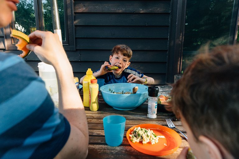 Boy is framed by two family members as he eats a pickle at the picnic table