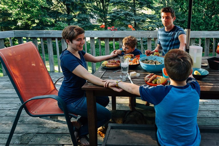 Family holds hands at dinner. Young boy is holding hands with his mother while trying to eat a pickle out of that hand.