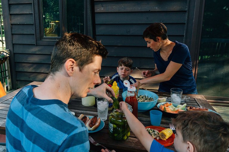 Family of four at outside picnic table. Dinner is on the table. Young son reaches for pickles while mother serves older son rice.