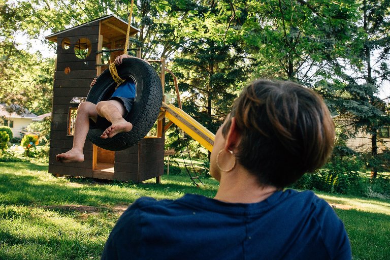 Boy swings on backyard tire swing in evening summer light while mother sits in grass and looks on