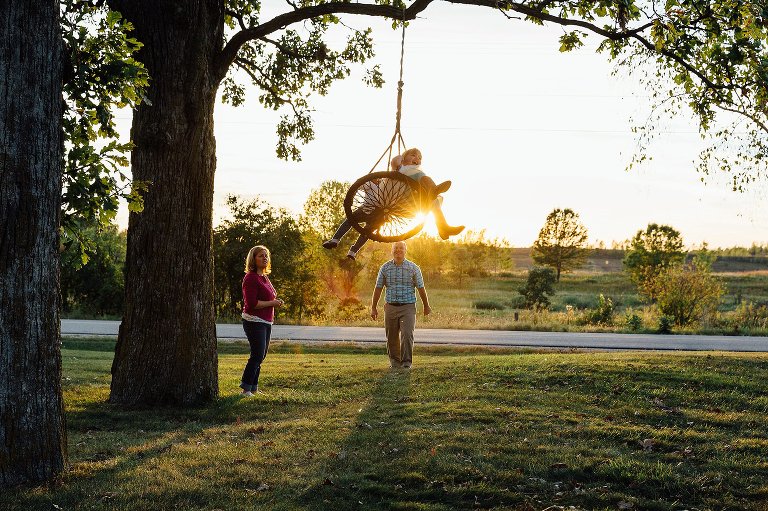 Mom and Dad push 2 daughter on the large swing under a tall oak tree at sunset. Sun flare comes through the swing. 