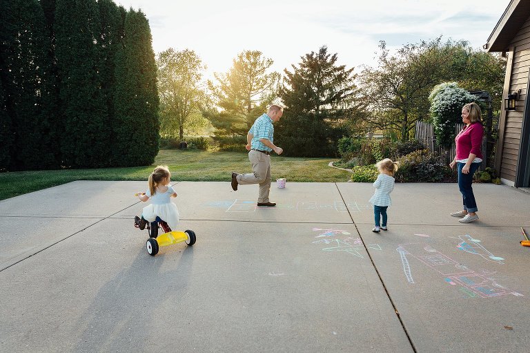 Dad hopscotches on the chalk hopscotch he made with his wife and two young daughters. The sun is low and soft behind him. 