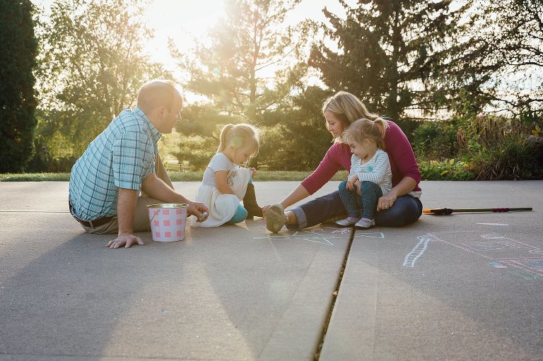 Mom, Dad, and two daughters make chalk creations in their driveway, The evening sun is soft, low, and warm. 