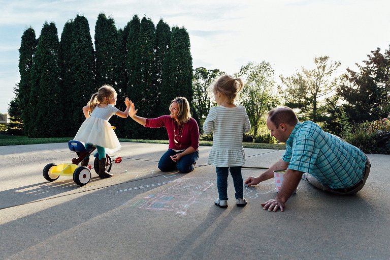 Family of four make chalk creations on their driveway. Mom hi-fives her older daughter on their work. 