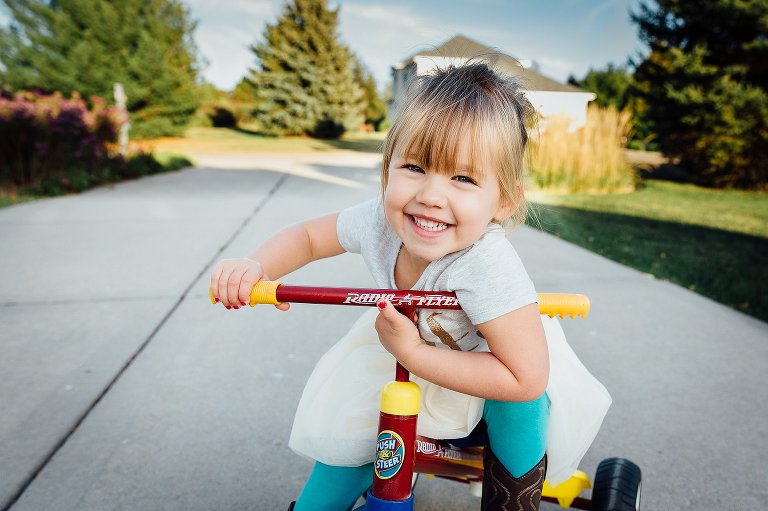 Four year old girl is smiling while leaning on her tricycle at the end of her long driveway. 