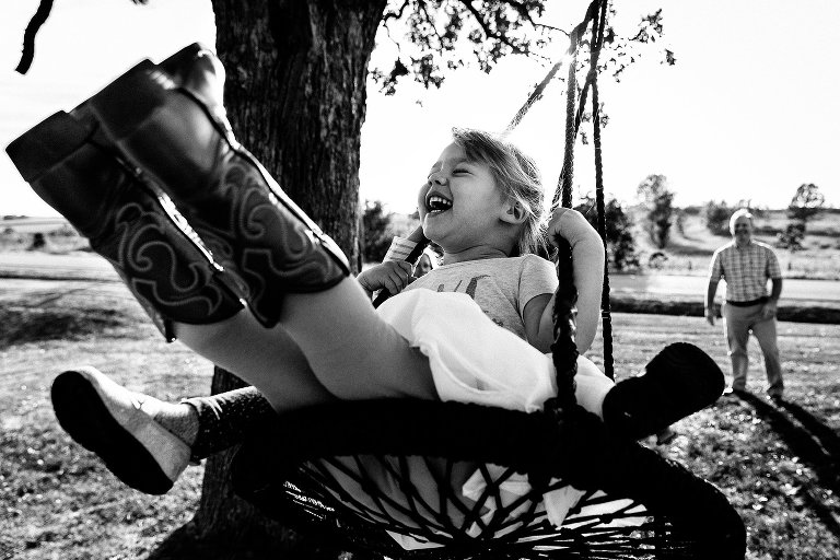 Black and white. Girl swings on a large swing, eyes. closed, and huge smile. Two small shoes are present behind her, indicating she is riding hte swing with a younger person behind her. 
