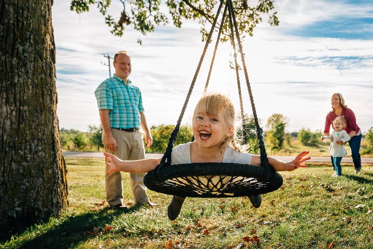 A young girl swings on a large swing under an oak tree. Her parents and sister wait and smile behind her. 