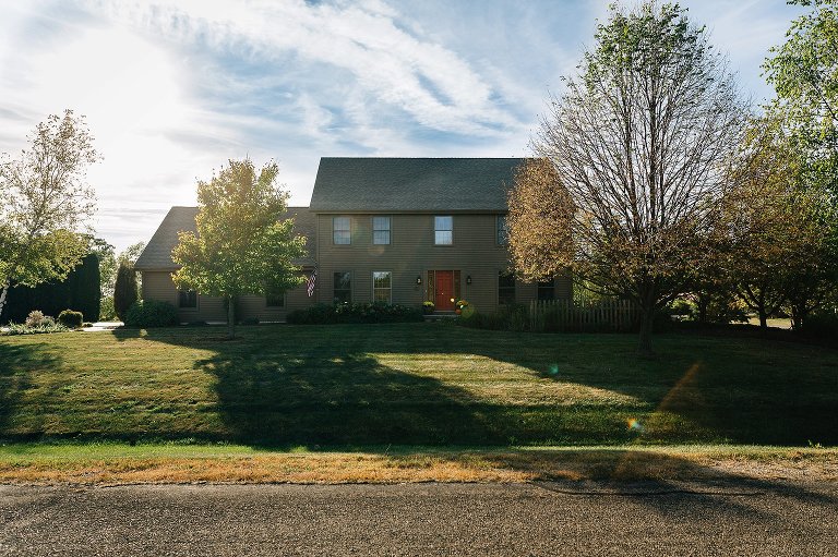 Big, beautiful brown house sits on large property in late fall. Blue skies and fallen leaves surround the scene. 