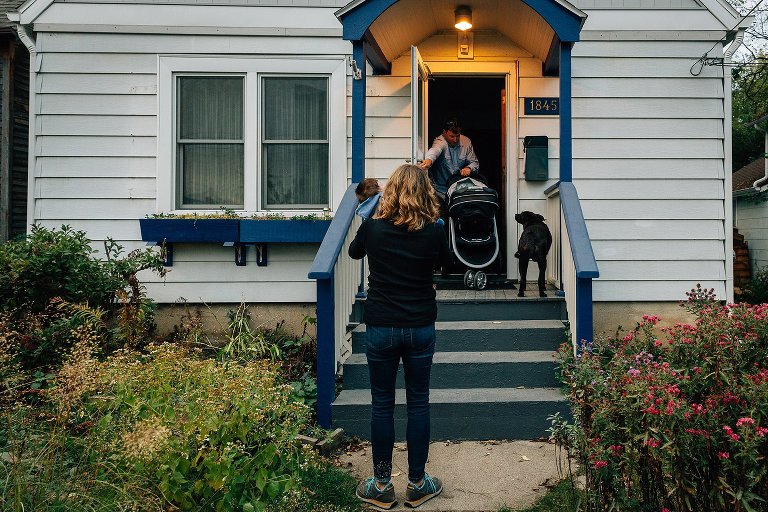 Dad holds door open as he tries to get large stroller through the door. Mom, baby and dog wait on the outside, watching. 