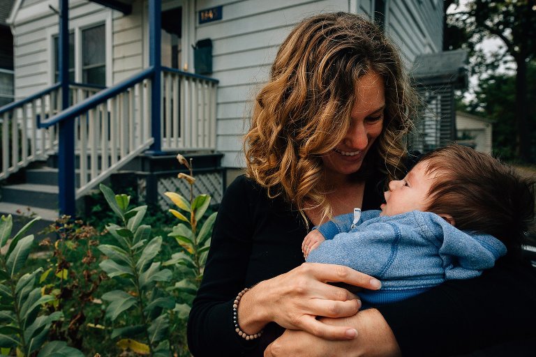 Mom smiles and holds infant daughter and smiles in front of home. 