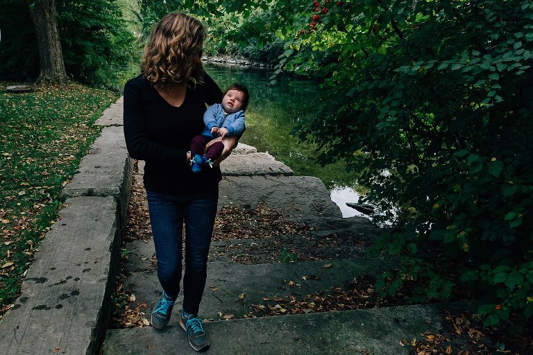 Dark shadows as mother walks away from a river holding infant daughter. Baby is looking up at sky, mom is looking at daughter. 