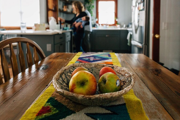 Focus on 4 yellow-red apples on dining room table. In the background, out of focus, mother holds sleeping baby and stirs pot on the stove. 