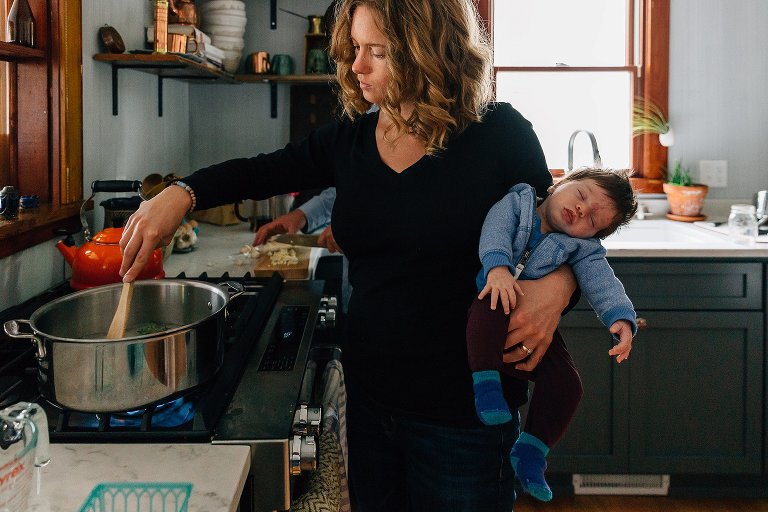 Baby is asleep in mother's arms as she stirs a pot on the stove with a wooden spoon. 