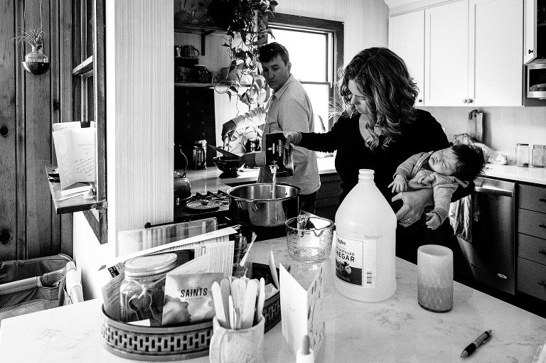 Mom holds baby with left hand, pours salt with right. Dad holds colander with left hand, tongs with right. A large container of vinegar is in the foreground. 