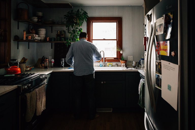 Man stands at kitchen sink. Back to the camera, illuminated by window light behind the sink. 