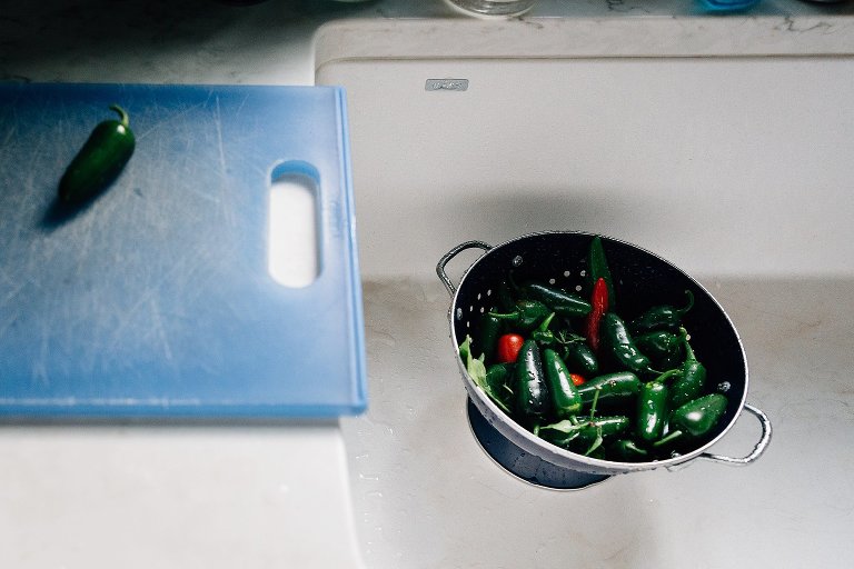 Freshly picked jalapenos are sitting in a colander in a white sink. Just washed. One green jalapeno lays on a blue cutting board. 