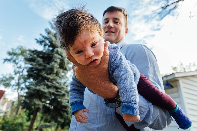 Dad holds baby on arm. View is looking up from ground at baby and dad and blue skies