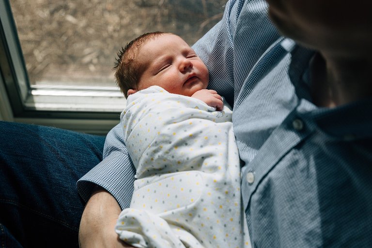 Dad holds sleeping, swaddled newborn in window light. 