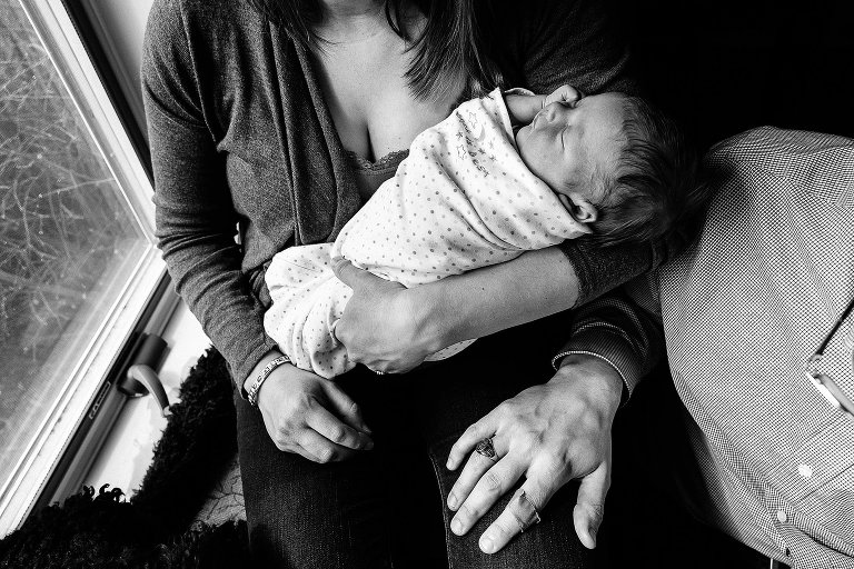 Black and white. Top down view. Mom holds sleeping, swaddled newborn while Dad places his hand on mom's leg. Dog paw in lower right corner. 