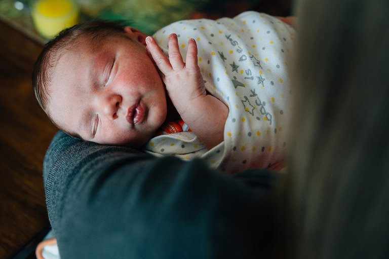 Top down view of newborn being held by mother. Newborn is sleeping and his hand has escaped the swaddle and we see three fingers near his face. 
