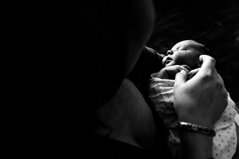 Black and white. Mother holds sleeping, swaddled newborn and touches his exposed fingers.