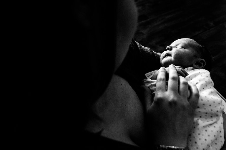 Black and white. Mother holds sleeping, swaddled newborn and touches his exposed hand.