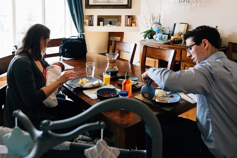 Parents eat breakfast while holding newborn. Pack n play in the foreground. 