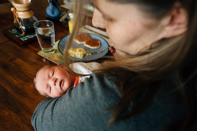 Mom holds newborn baby at the breakfast table. Pancakes, eggs, and French press coffee on the table. 