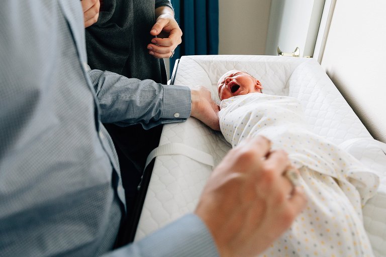 Swaddled newborn yawn-cries after being changed on changing table. Both parents are present and hands are in view. 