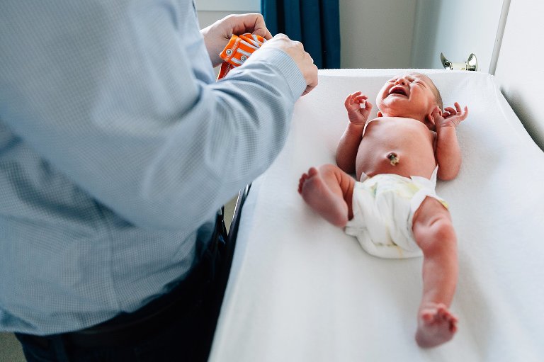 Dad preparing to change newborn clothes. Attached umbilical cord on belly button. Baby is crying, leg stretched out. 