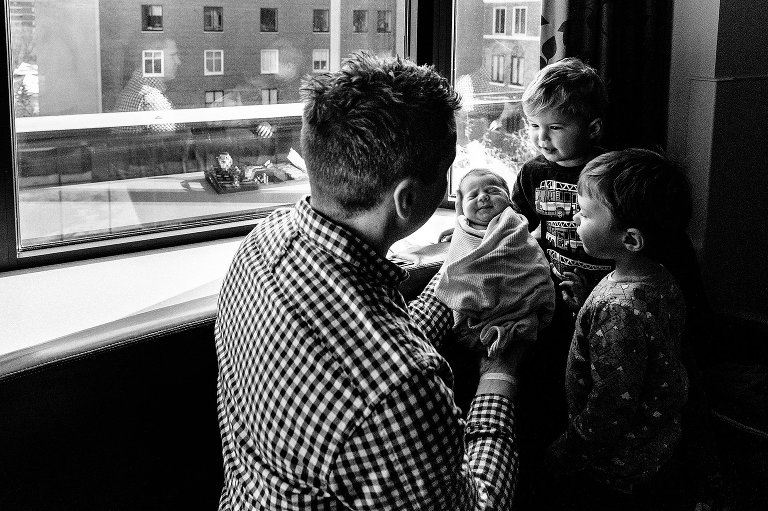 Black and white. Dad holds up newborn daughter for his two older brothers. Location: hospital room after baby is born. 