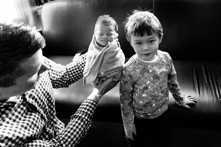 Black and white. Dad holds up newborn sister to compare how big the older sibling is. 