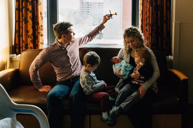 Family of five sits on the couch at hospital. Dad flies a Lego helicopter over the family. 