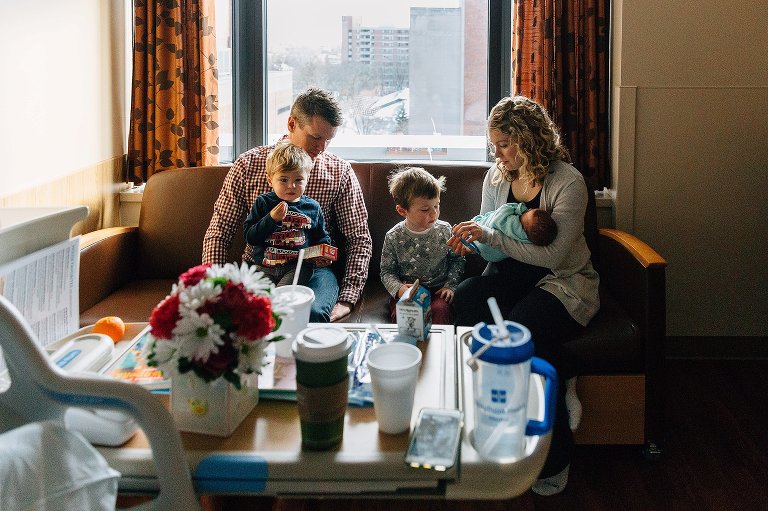 Family of five sit on couch in hospital room after newborn was born. 