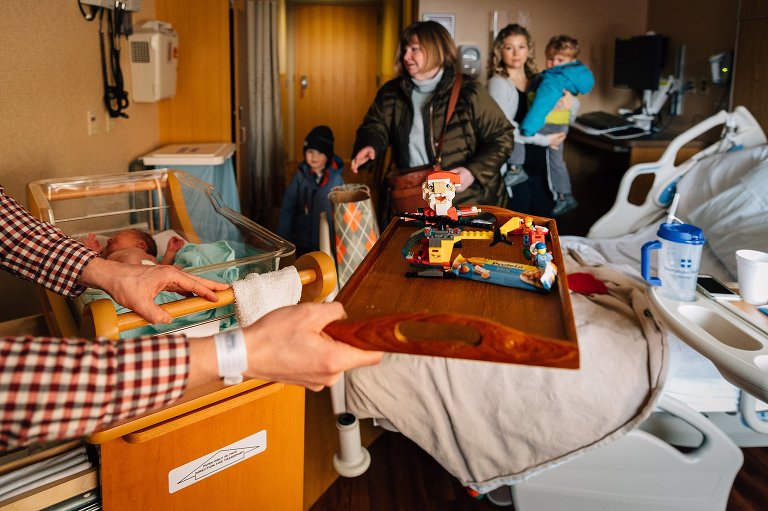 Dad holds a tray of built Santa Legos as grandparents and older siblings enter hosptial room to meet newborn sister. 