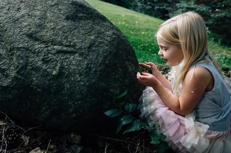 A young girl crouches behind a large gray boulder and discovers a flower and examines it closely.