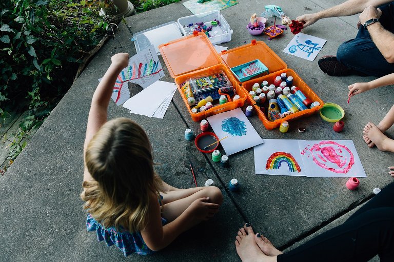 Top down view of girl painting on back porch. There are paints organized in tubs and vibrant illustrations on the ground.