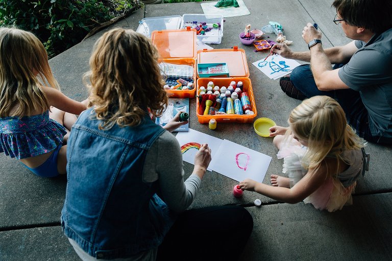 Top down view. Family of four sits on back patio and paints pictures together. Painted papers are scattered and the family is engaged with each other.