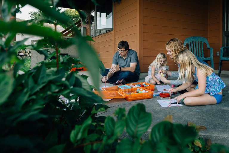 Family of four sits on back patio and paints pictures together. Painted papers are scattered and the family is engaged with each other.