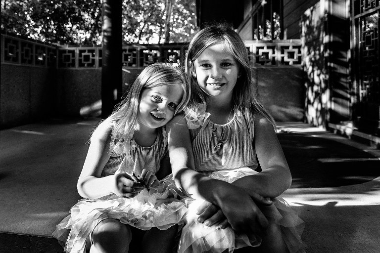 Black and white. Portrait of two girls sitting on their front steps. Dappled light, smiling, camera aware.