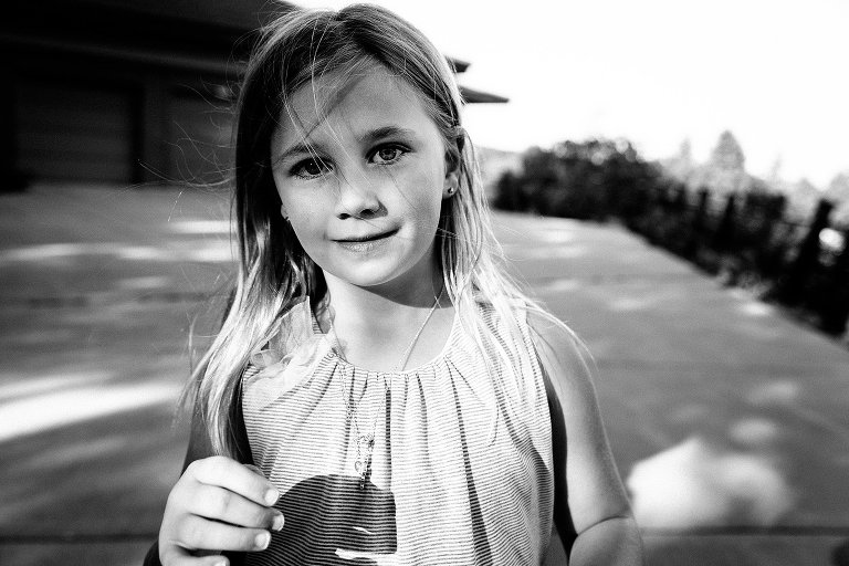 Black and white. Young girl is smiling, camera aware on her driveway. There is late day dappled light and long shadows.