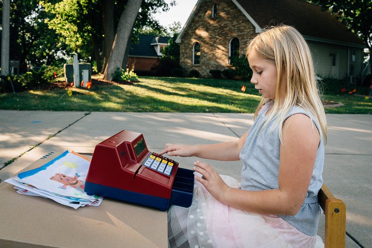 Girl sits at her cardboard box table and cash register and waits for someone to come buy one of her watercolor illustrations.
