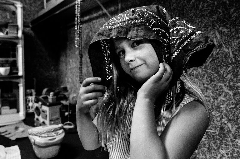 Black and white. Girl puts handkerchief on her head in her dress-up/role play play. She is slightly smiling and camera aware.