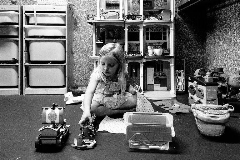 Young girl lays out toys in front of her and sits on the basement floor toyroom to play. She reaches out with her right hand to grab a toy. Black and white.