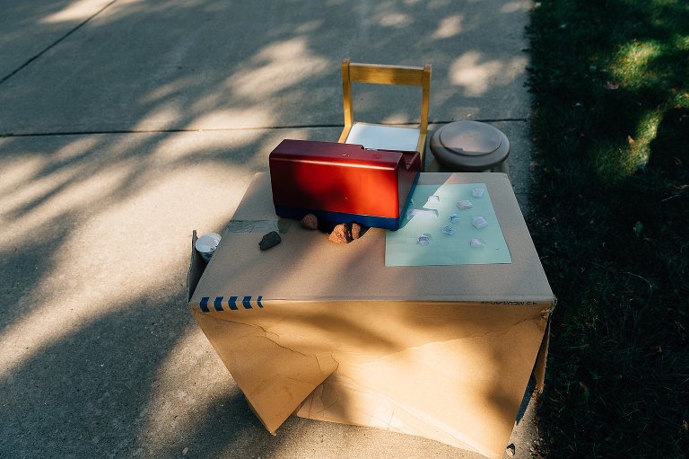 A cardboard box and toy cash register sits in the driveway under dappled summer light. This is a sales stand for a young child.