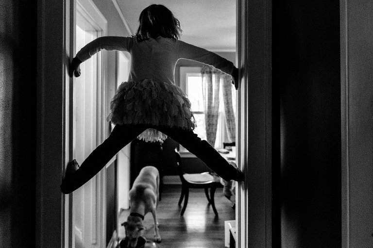 Girl climbs the space in the doorway. Black & white. Family dog get a drink under her. 