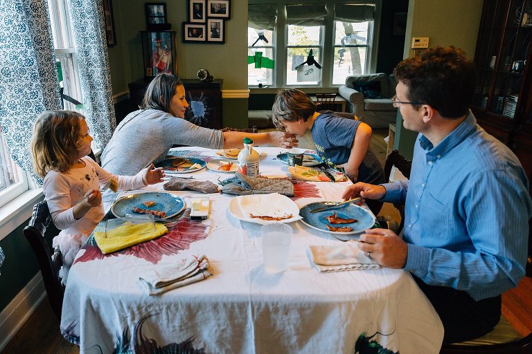 Mom helps son bite off bacon in his mouth while the rest of the famliy at the breakfast and watches. 