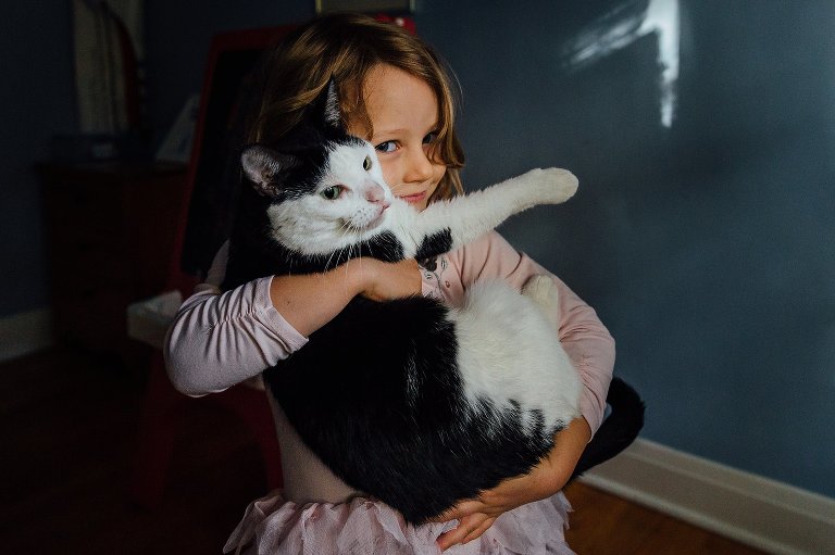 Young girl holds a cat in window light. She makes eye contact with camera and slightly smiles.