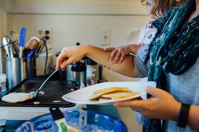Daughter holds onto to mom's arm while mom flips pancakes wiht one hand and holds a plate of pancakes with the other. 