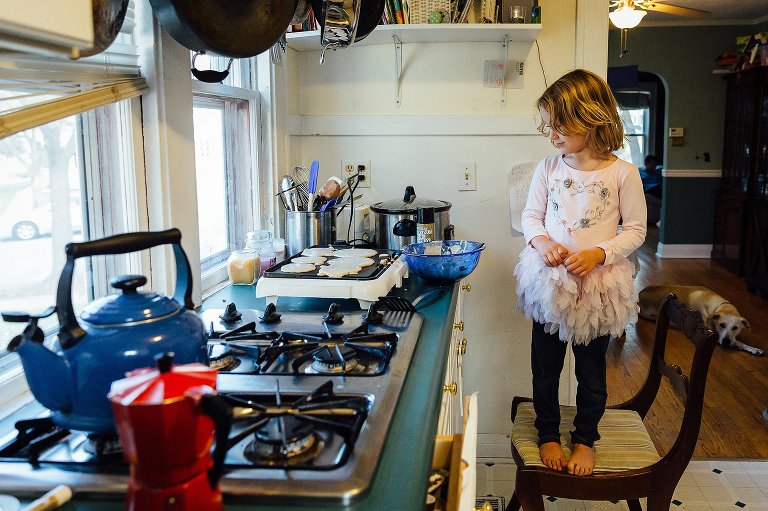 Portrait of a young girl standing on a chair in the kitchen, looking at pancakes on the electric griddle. 